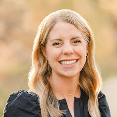 A blonde haired woman in a dark shirt smiles at the camera.